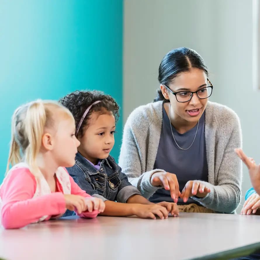 Kita Stageteaser Eine junge Erzieherin sitzt mit einer Kindergruppe an einem Tisch, ein kleiner Junge macht mit Gesten auf sich aufmerksam