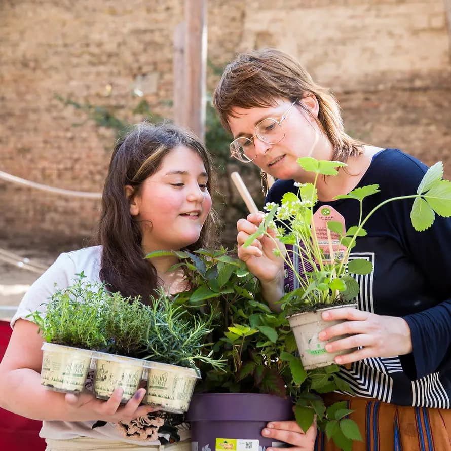 94414_ SOS-KD_Elly_Berlin_Magazin_SebastianPfuetze_30.4.2024_1747.jpg Julia Willenborg, Mitarbeiterin bei SOS-Kinderdorf, und Mädchen halten Kräutertöpfe in den Händen