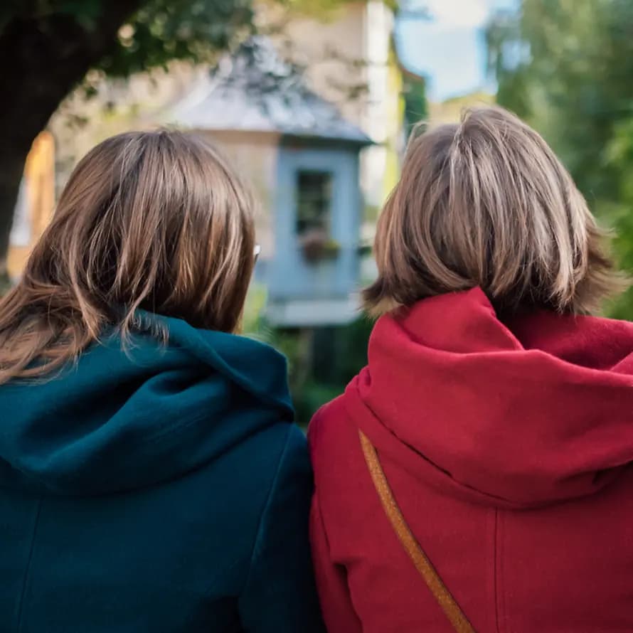 Beitrag Geschwisterlichkeit Zwei Frauen mit hellbraunem, mittellangem Haar sitzen Seite an Seite mit dem Rücken zum Betrachter