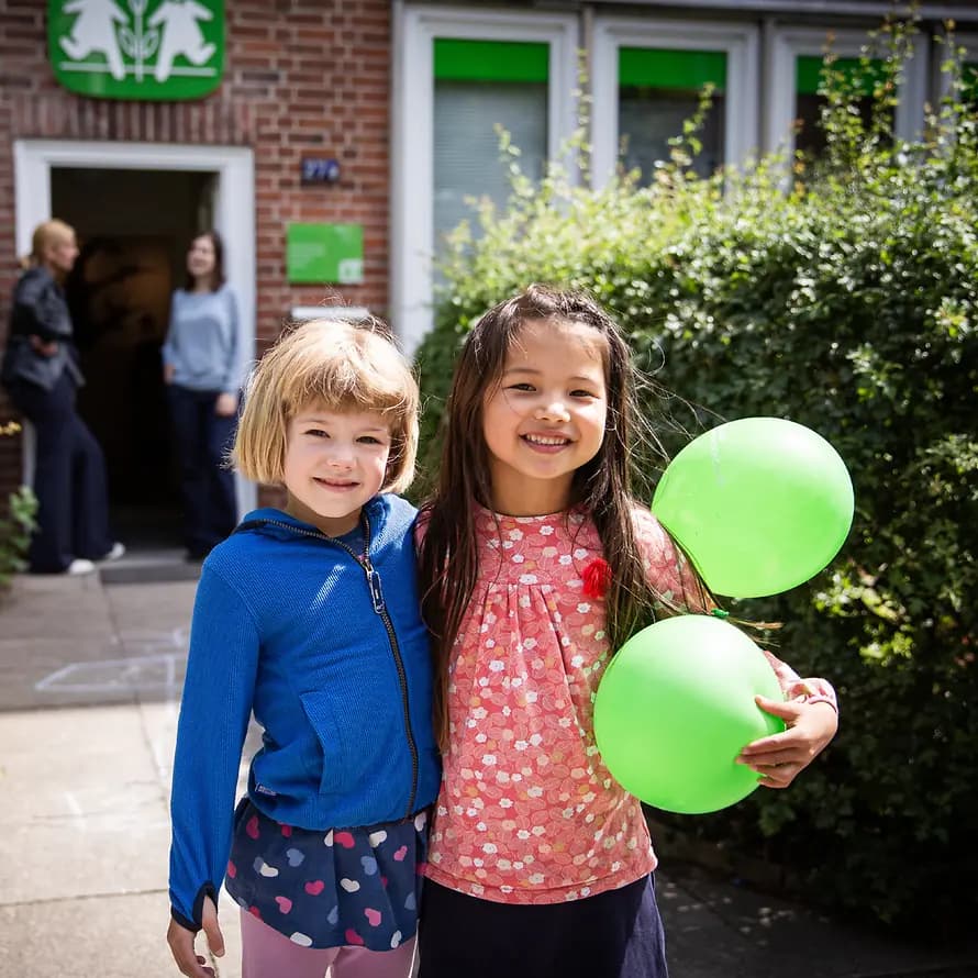 SOS-Kinderdorf hilft Kindern und Jugendlichen Zwei Mädchen mit grünen Luftballons vor einem Backsteingebäude von SOS-Kinderdorf